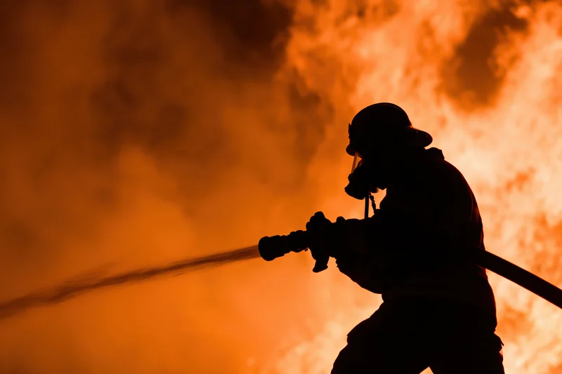 Firefighter silhouetted holding firehose against orange flames and smoke