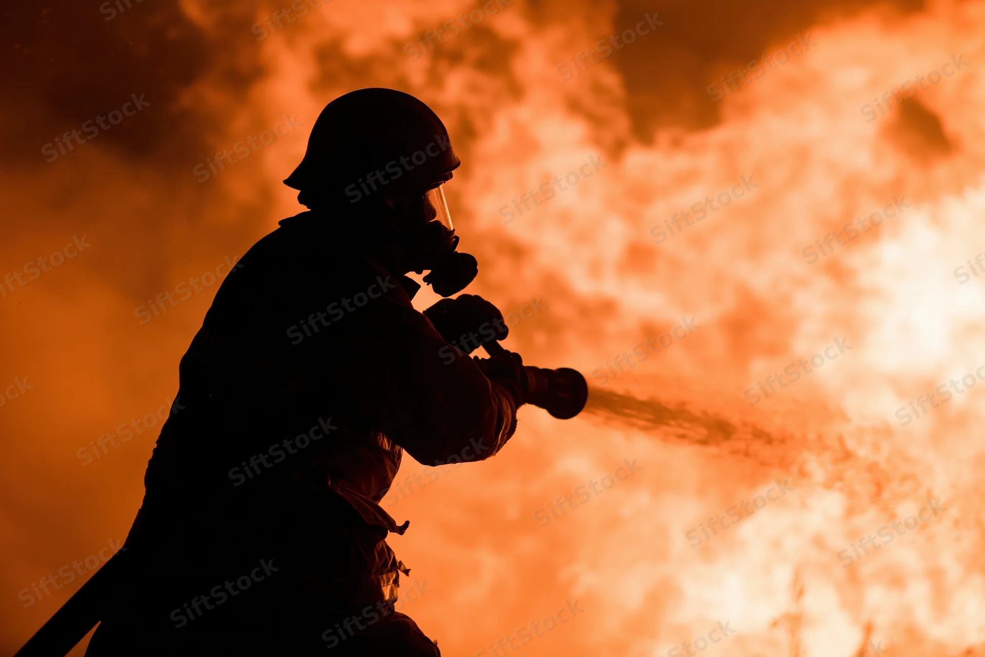 Firefighter in silhouette spraying water from hose against intense orange flames