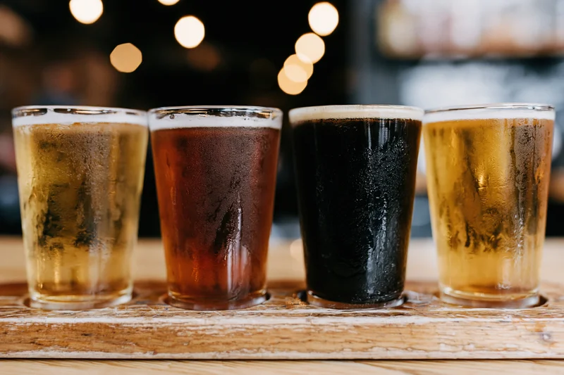 Four craft beer tasting glasses in a row on a wooden tray with condensation and foam, set against warm ambient lighting with bokeh background