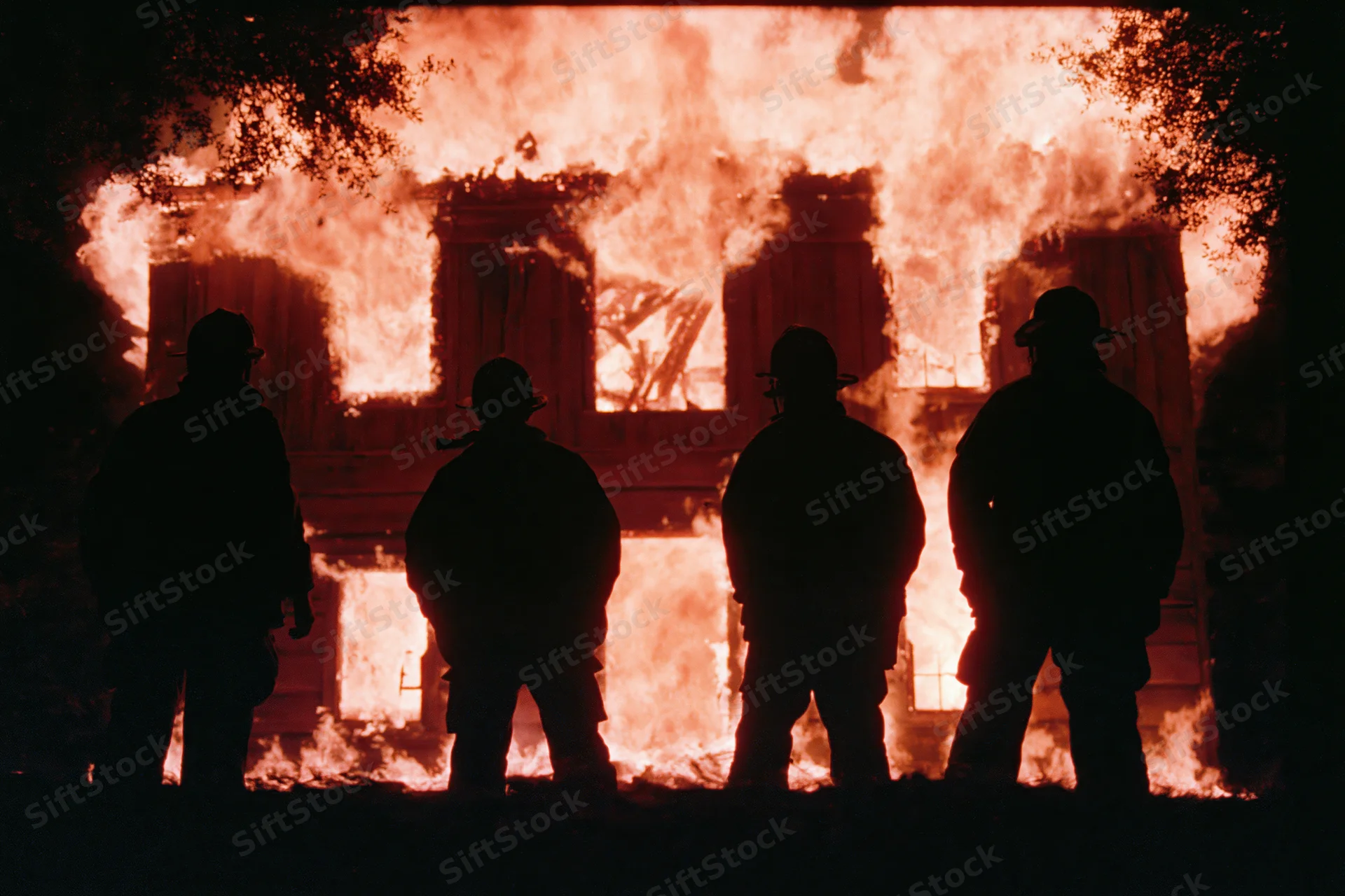 Four firefighters silhouetted against a burning two-story wooden building at night