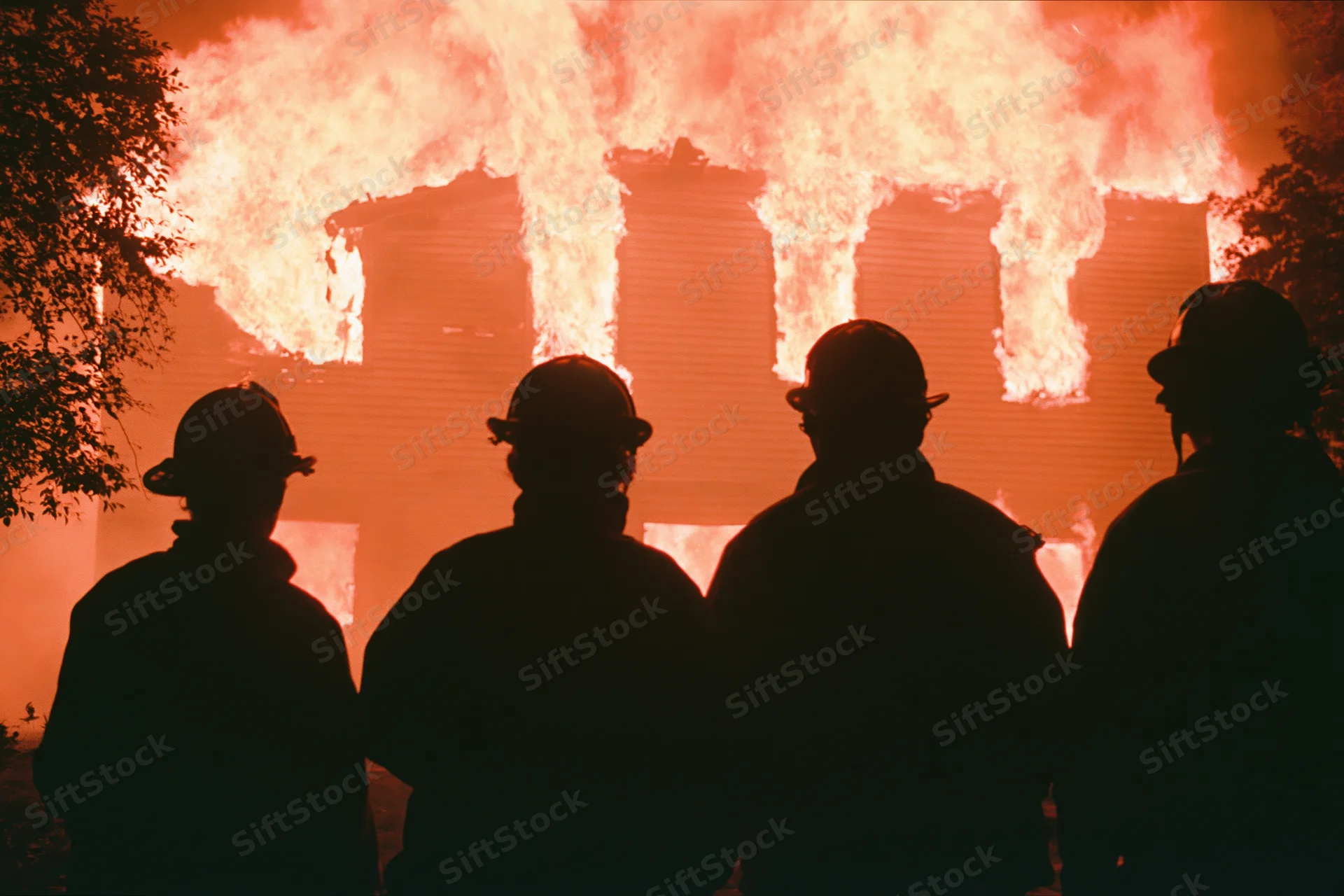 Four firefighters in silhouette facing a burning two-story house at night
