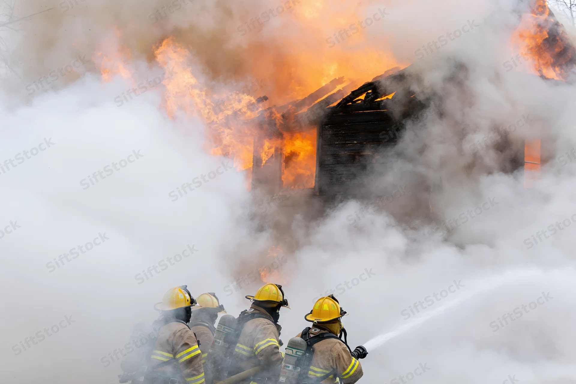 Firefighters in turnout gear spraying water at a burning house through thick smoke