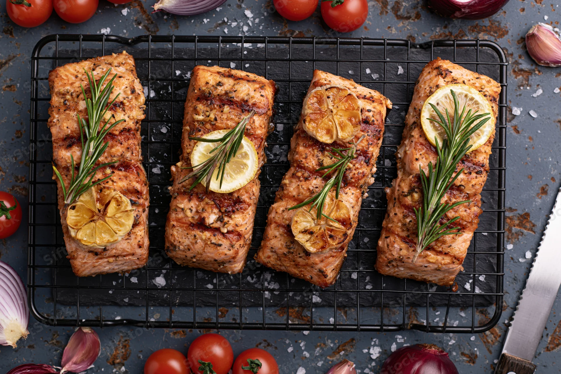 Four grilled salmon fillets garnished with lemon and rosemary on a wire rack surrounded by tomatoes and onions