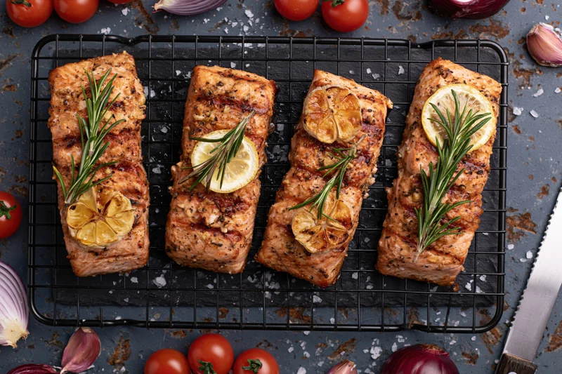 Four grilled salmon fillets garnished with lemon and rosemary on a wire rack surrounded by tomatoes and onions