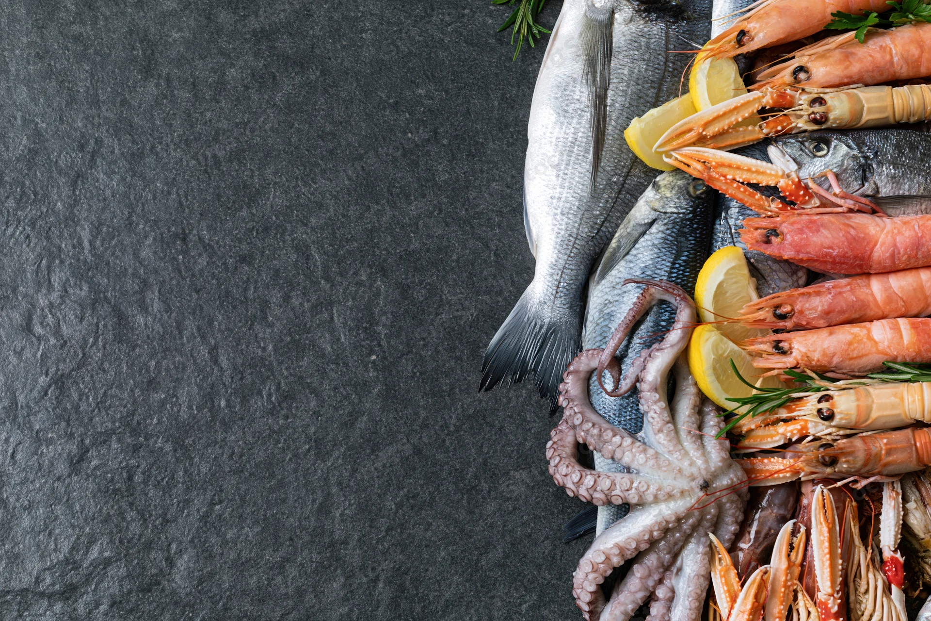 Fresh whole fish octopus langoustines and mixed seafood arranged on dark slate surface with lemon and herbs