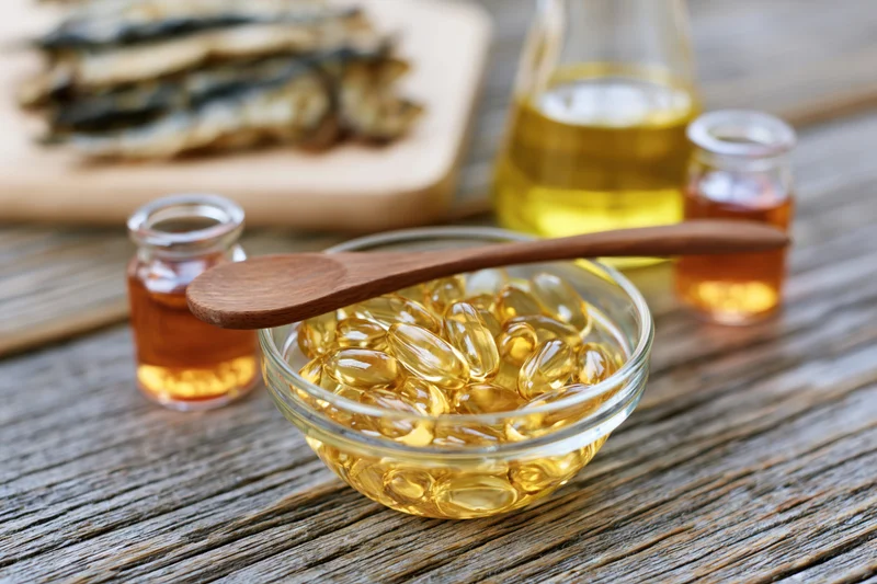 Glass bowl filled with golden omega-3 fish oil gel capsules on rustic wooden table with bottles and dried fish in background