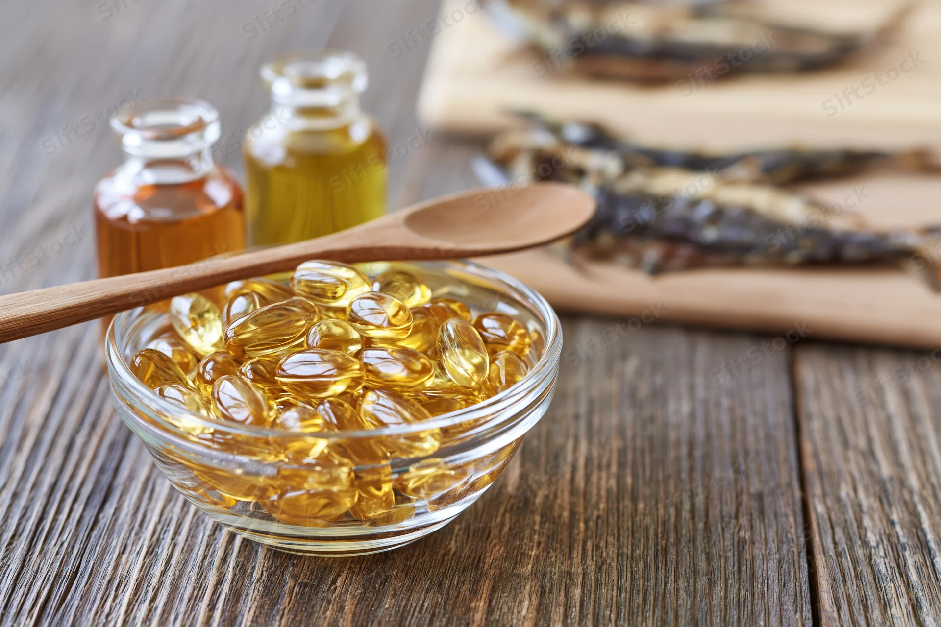Glass bowl filled with golden omega-3 fish oil gel capsules on wooden table with wooden spoon and bottles of oil in background