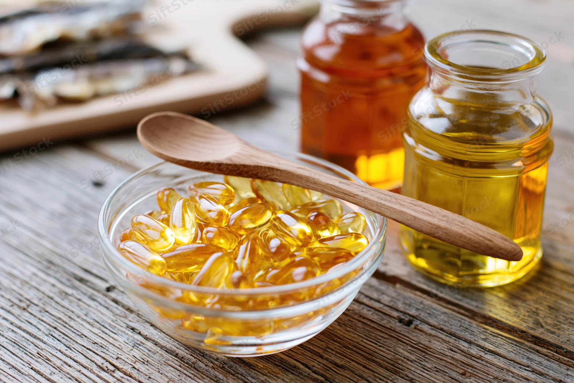 Glass bowl filled with golden omega-3 fish oil gel capsules on wooden table with jars of oil and wooden spoon