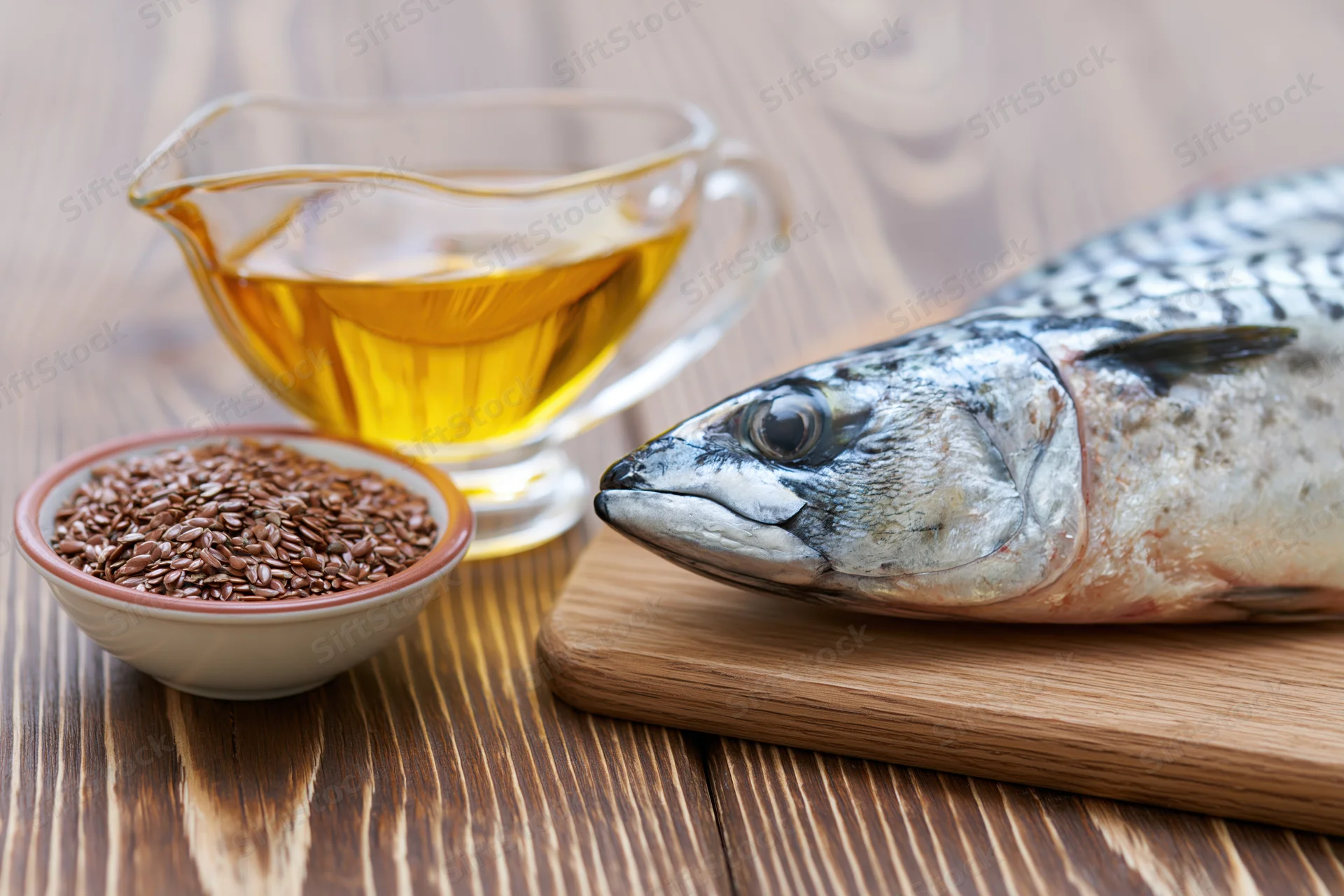 Whole fish on a wooden cutting board beside a glass gravy boat filled with golden oil and a bowl of flaxseeds on a wooden table