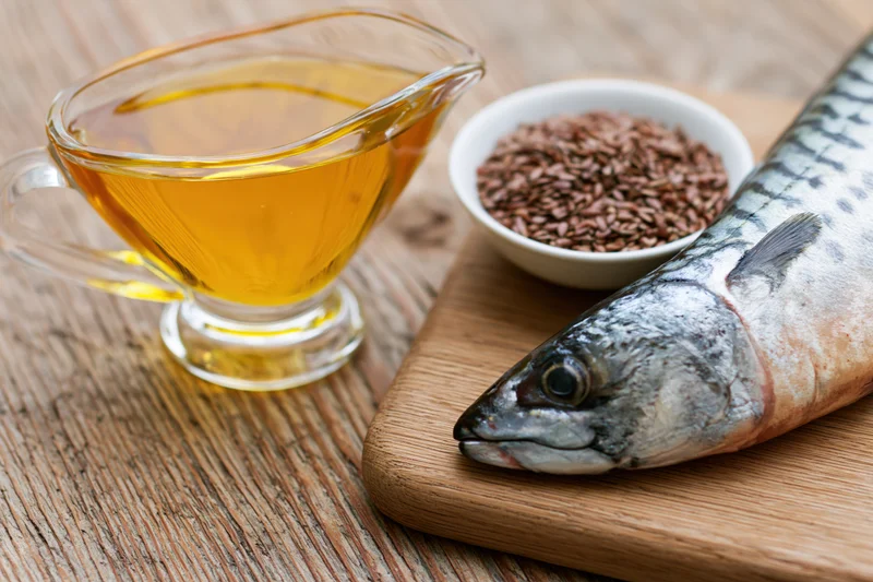 Raw mackerel fish on wooden cutting board beside glass gravy boat filled with golden fish oil and small bowl of flaxseeds