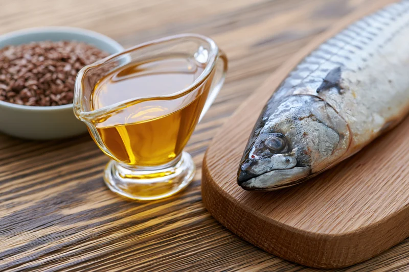 Whole fish on wooden cutting board beside glass gravy boat filled with golden oil and bowl of flaxseeds on wooden table
