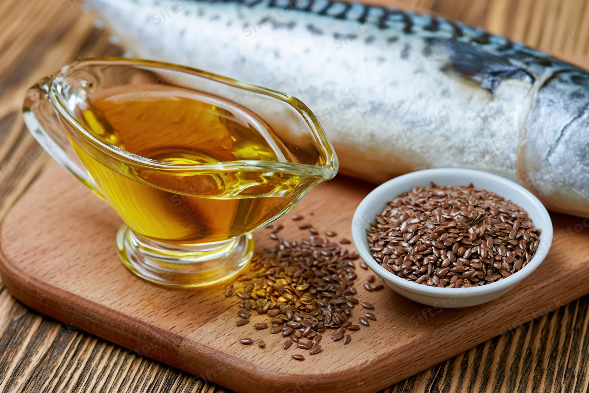 Glass gravy boat filled with golden fish oil beside flaxseeds and a whole fish on a wooden cutting board