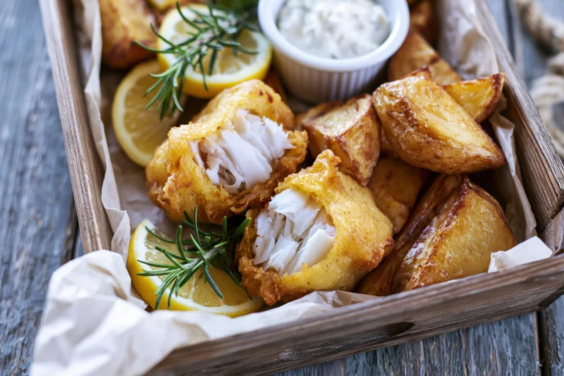 Golden battered fish fillet broken open to reveal flaky white interior served with roasted potato wedges and lemon slices in a wooden tray lined with parchment paper