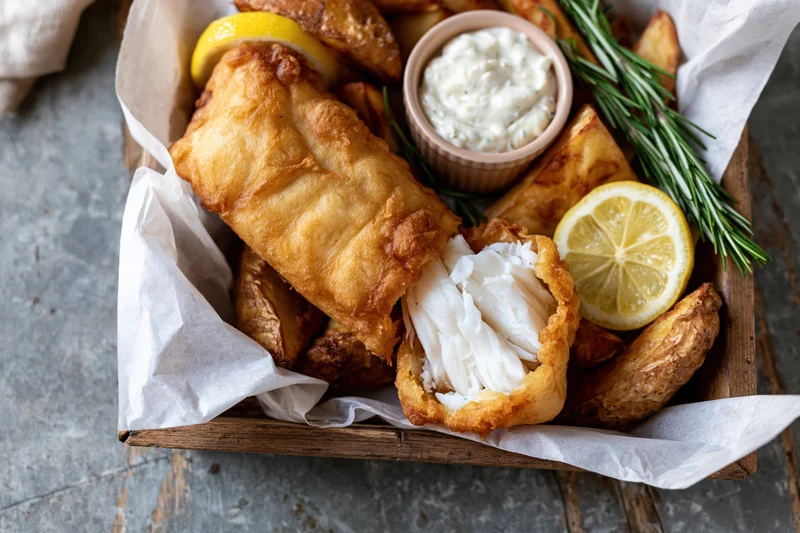 Golden battered fish fillet broken open to reveal flaky white interior served with chips lemon and tartar sauce in a wooden tray lined with parchment paper
