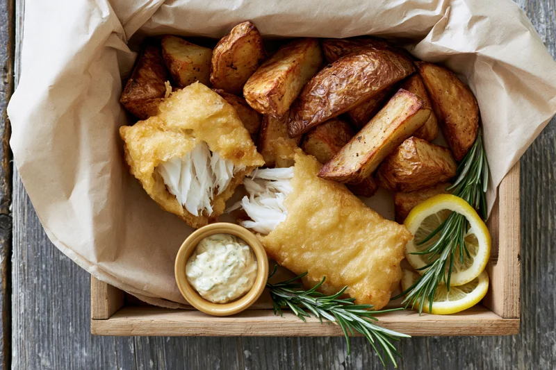 Golden battered fish fillet broken open with roasted potatoes and tartar sauce in a wooden tray lined with paper