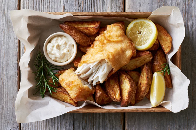 Golden battered fish fillet broken open to reveal flaky white meat with potato wedges, lemon, rosemary, and tartar sauce in a wooden tray lined with parchment paper on a gray wooden surface