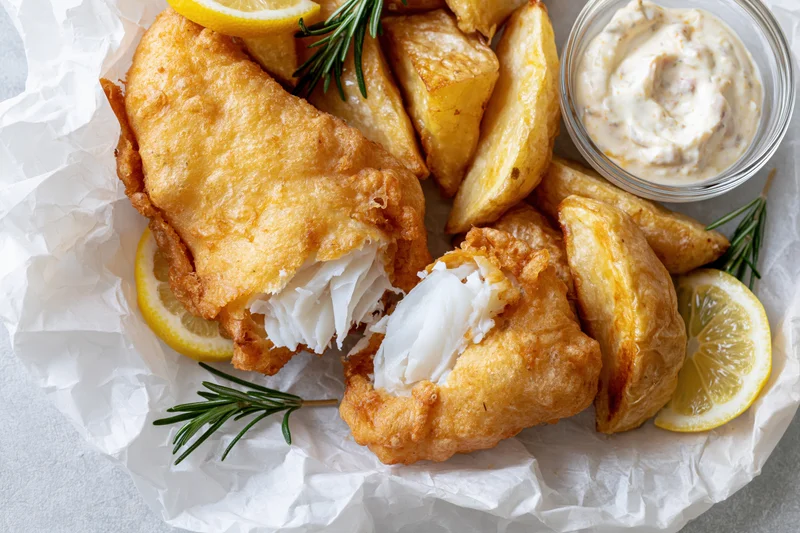 Golden battered fish fillet with broken piece revealing white flaky interior served with potato wedges lemon slices rosemary and tartar sauce on crumpled white paper