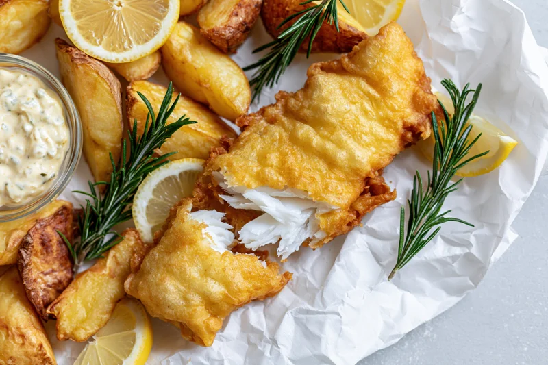 Golden battered fish fillet with broken piece revealing white flaky interior served with lemon slices, rosemary, chips and tartar sauce on crumpled white paper