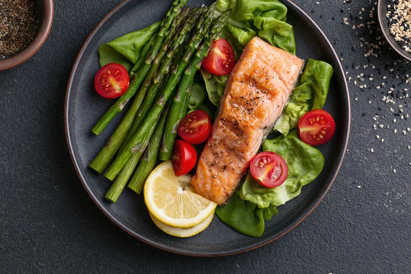 Grilled salmon fillet with asparagus and cherry tomatoes on a bed of lettuce on a dark plate