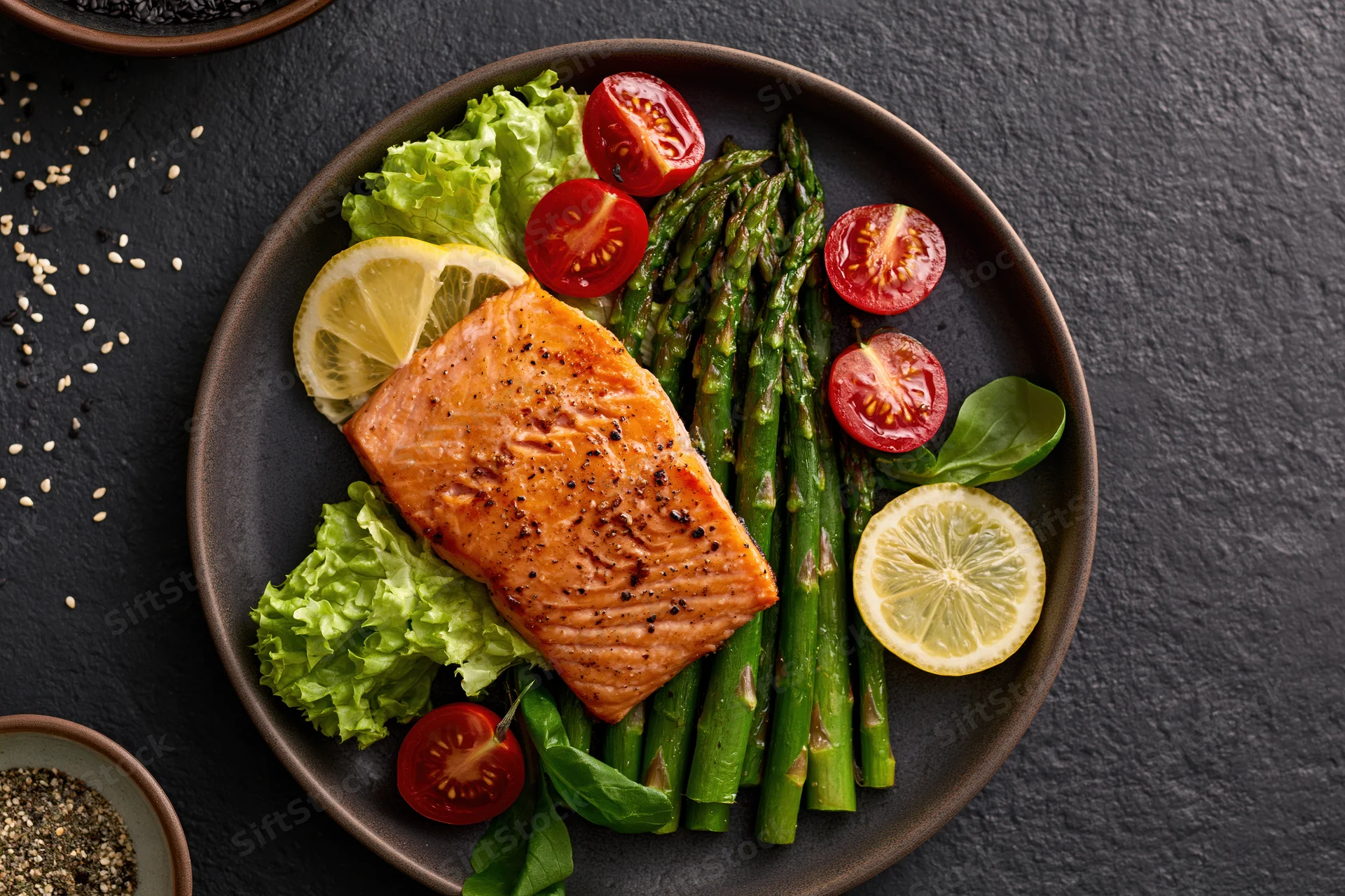 Grilled salmon fillet with asparagus and cherry tomatoes on a dark plate with lemon and lettuce