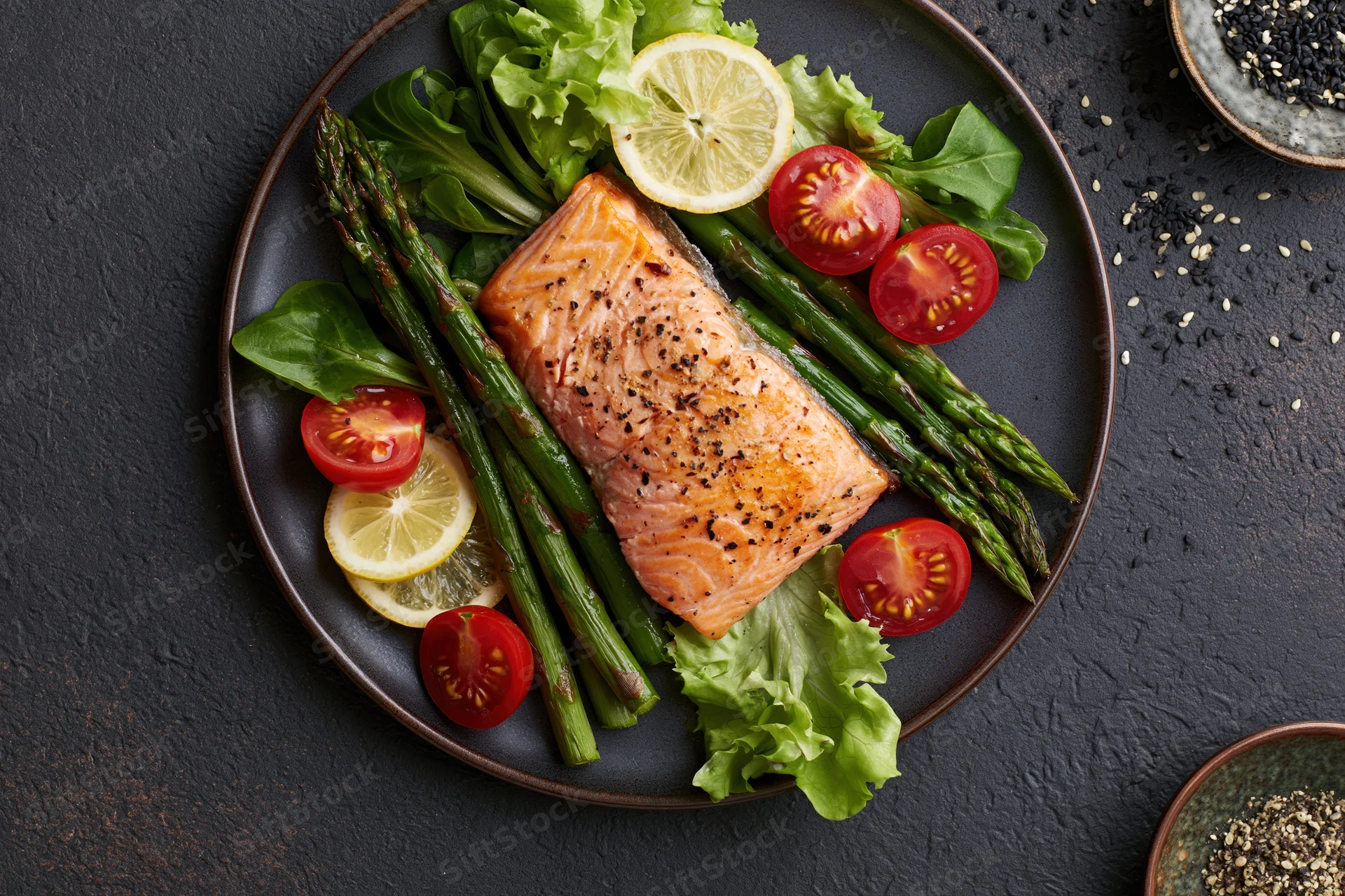 Grilled salmon fillet with asparagus and cherry tomatoes on a dark plate with lemon and lettuce garnish
