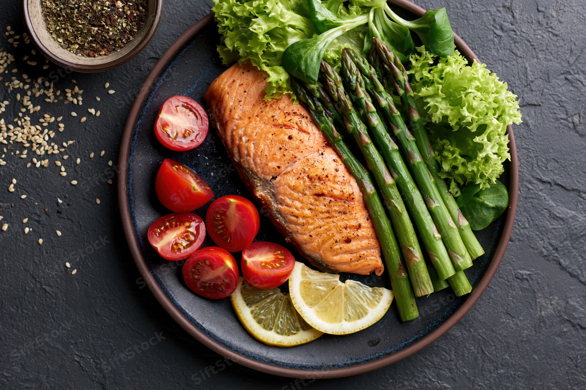 Grilled salmon fillet with asparagus and cherry tomatoes on a dark plate with lemon and lettuce