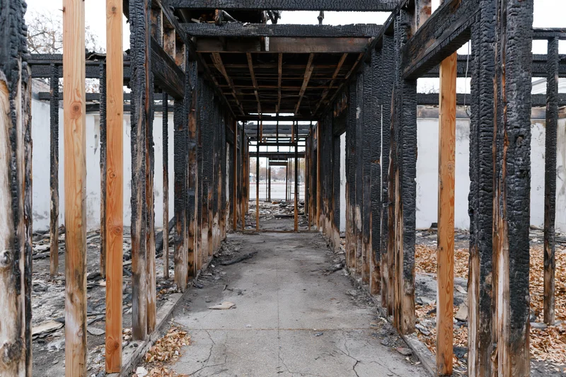 Charred wooden frame of a fire-damaged building interior with exposed beams and debris on concrete floor