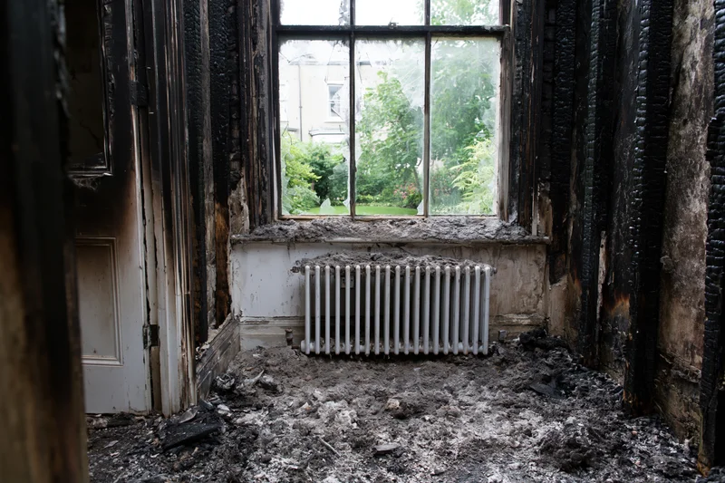 Fire-damaged room interior with charred walls and debris around a radiator under a window