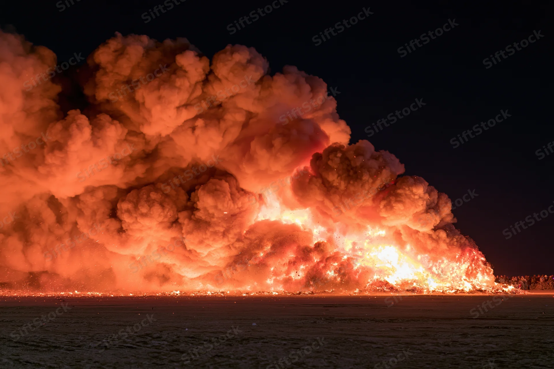 Massive fire burning across a forested landscape at night, producing thick orange smoke and intense flames under a dark sky