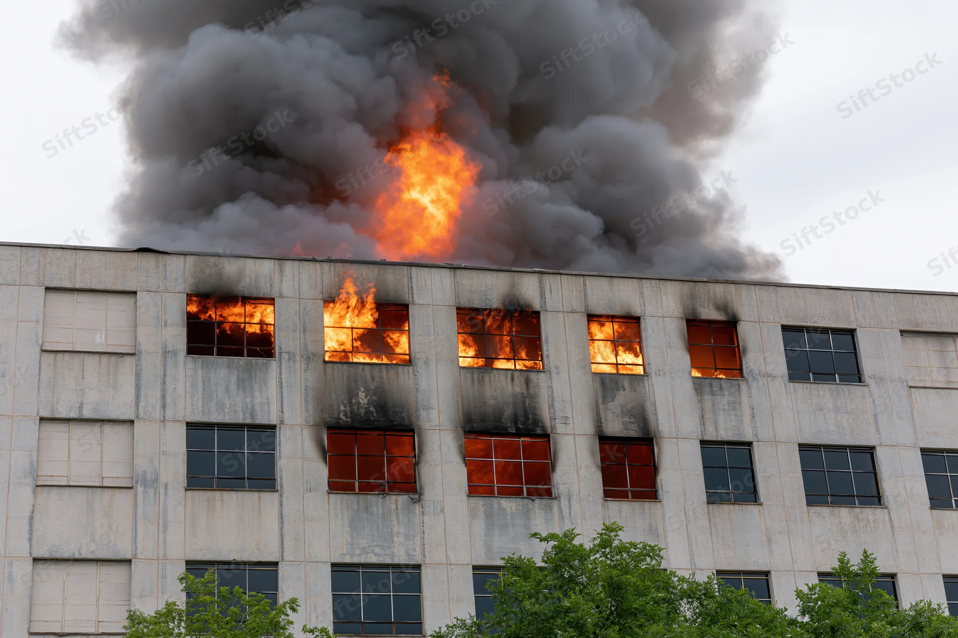 Large industrial concrete building engulfed in flames with thick smoke billowing from windows