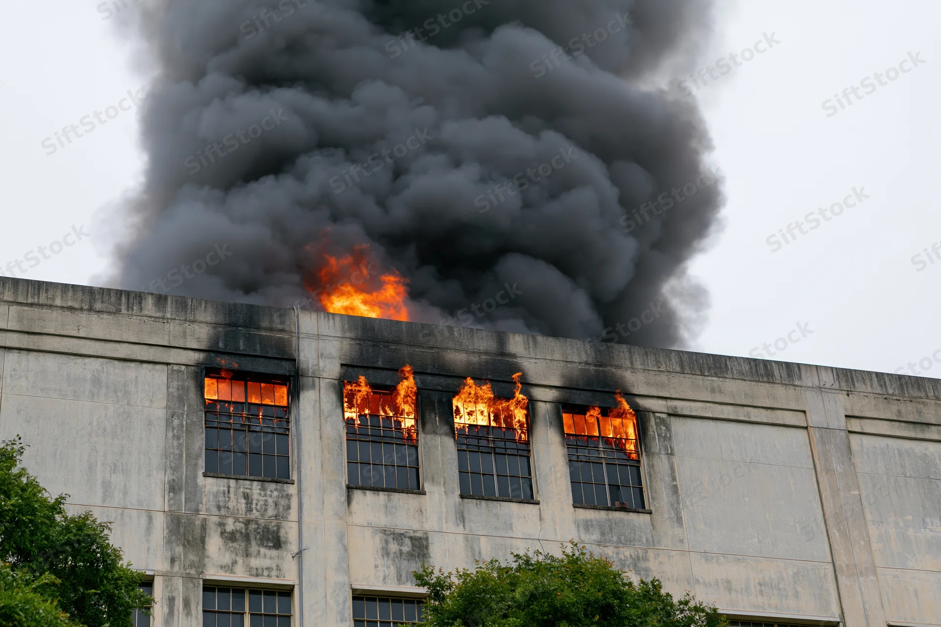 Flames erupting from windows of a large industrial concrete building with thick black smoke rising above