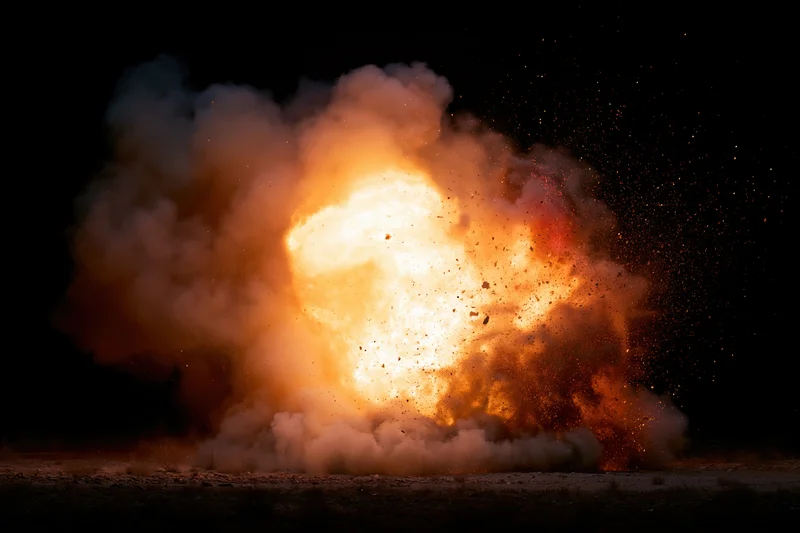 Large explosion bursting at ground level at night with billowing smoke and flying debris