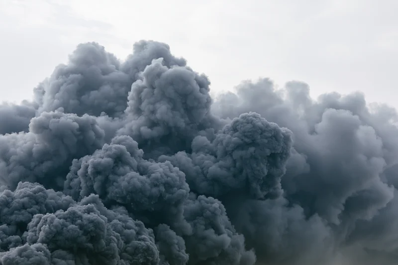 Massive billowing column of dark grey smoke rising into the overcast sky