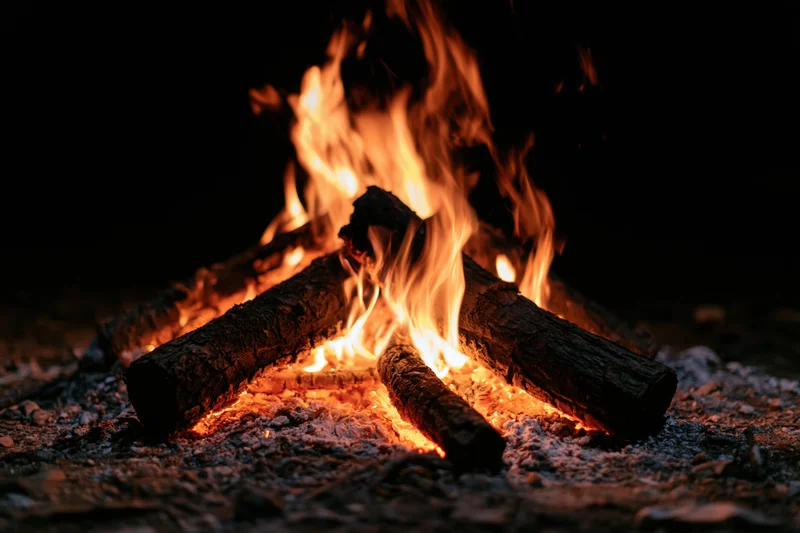 Campfire burning with flames rising from stacked logs on dark ground at night