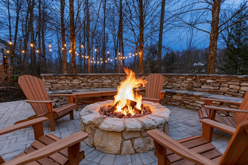 Stone firepit with flames surrounded by adirondack chairs on a brick paver patio at twilight