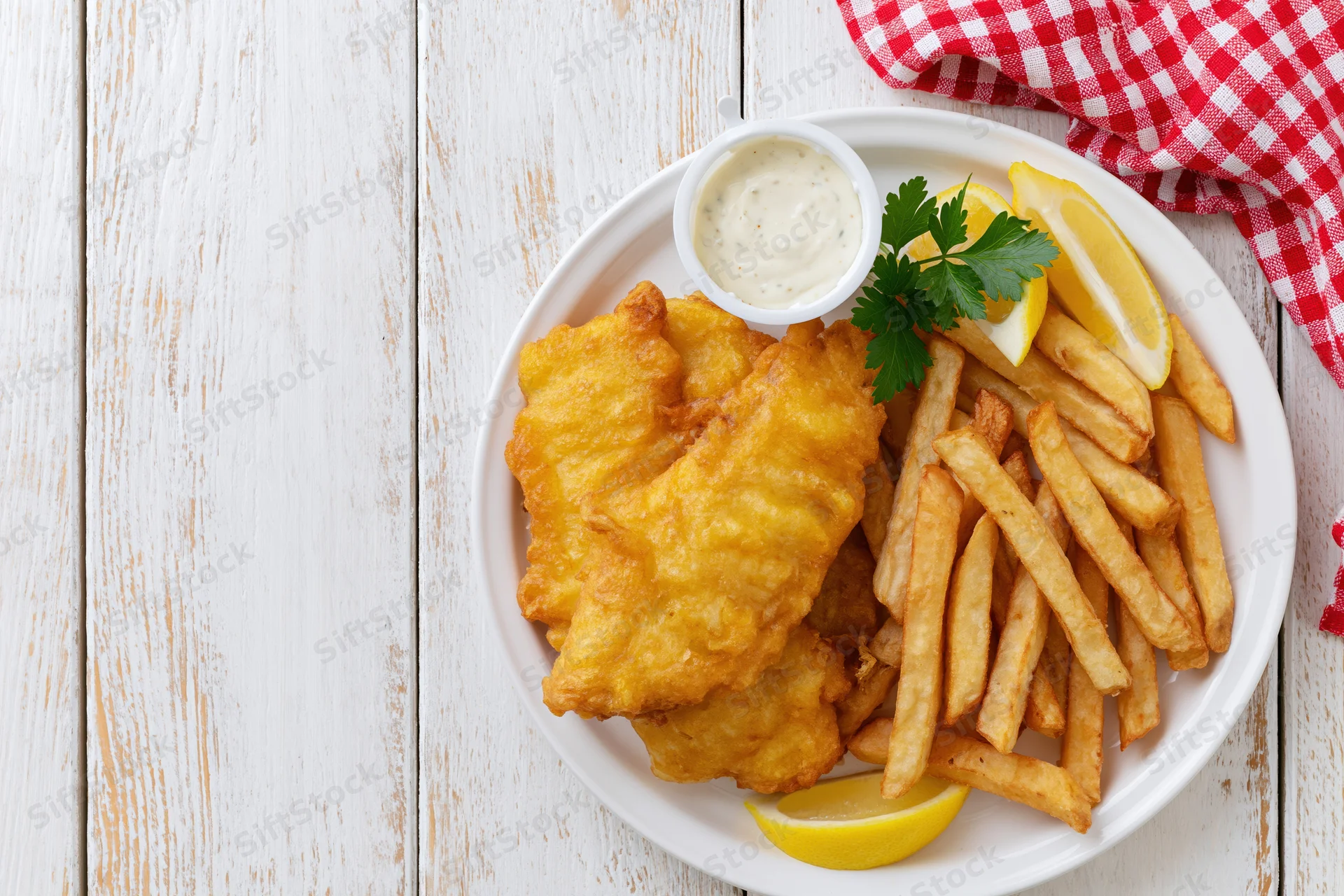 Golden battered fish and french fries on a white plate with lemon and tartar sauce, set on a rustic wooden table with a red checkered napkin
