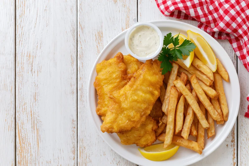 Golden battered fish and french fries on a white plate with lemon and tartar sauce, set on a rustic wooden table with a red checkered napkin