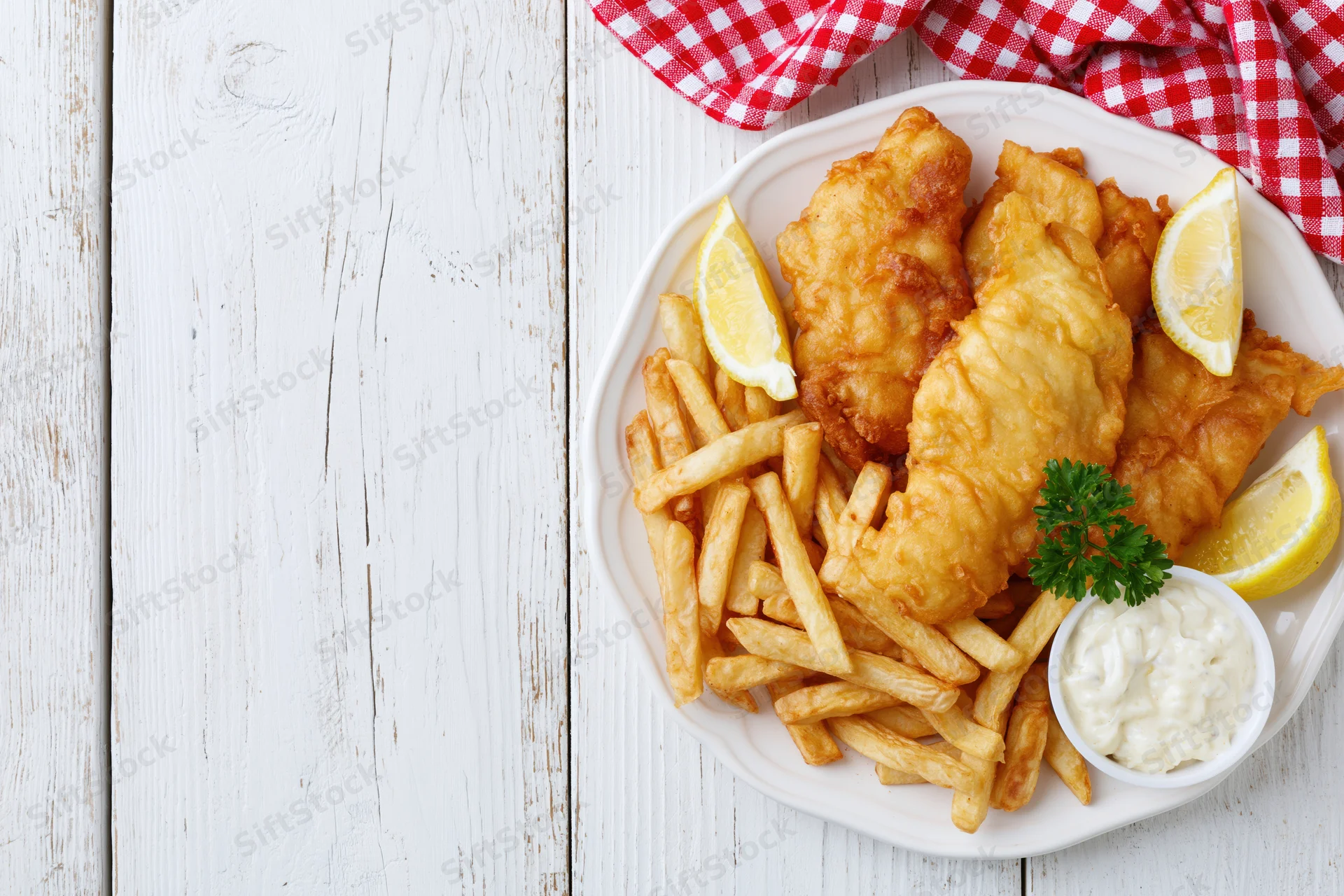 Fried fish and french fries on a white plate with lemon wedges and tartar sauce, set on a white wooden table with a red checkered napkin