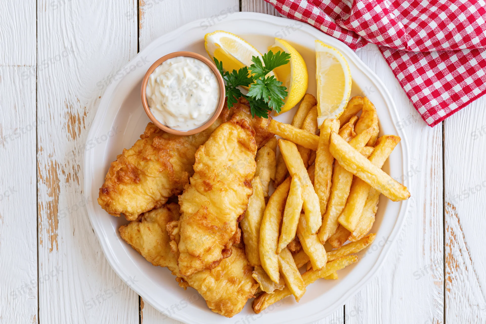 Golden battered fish and french fries on a white plate with tartar sauce and lemon garnish on a wooden table
