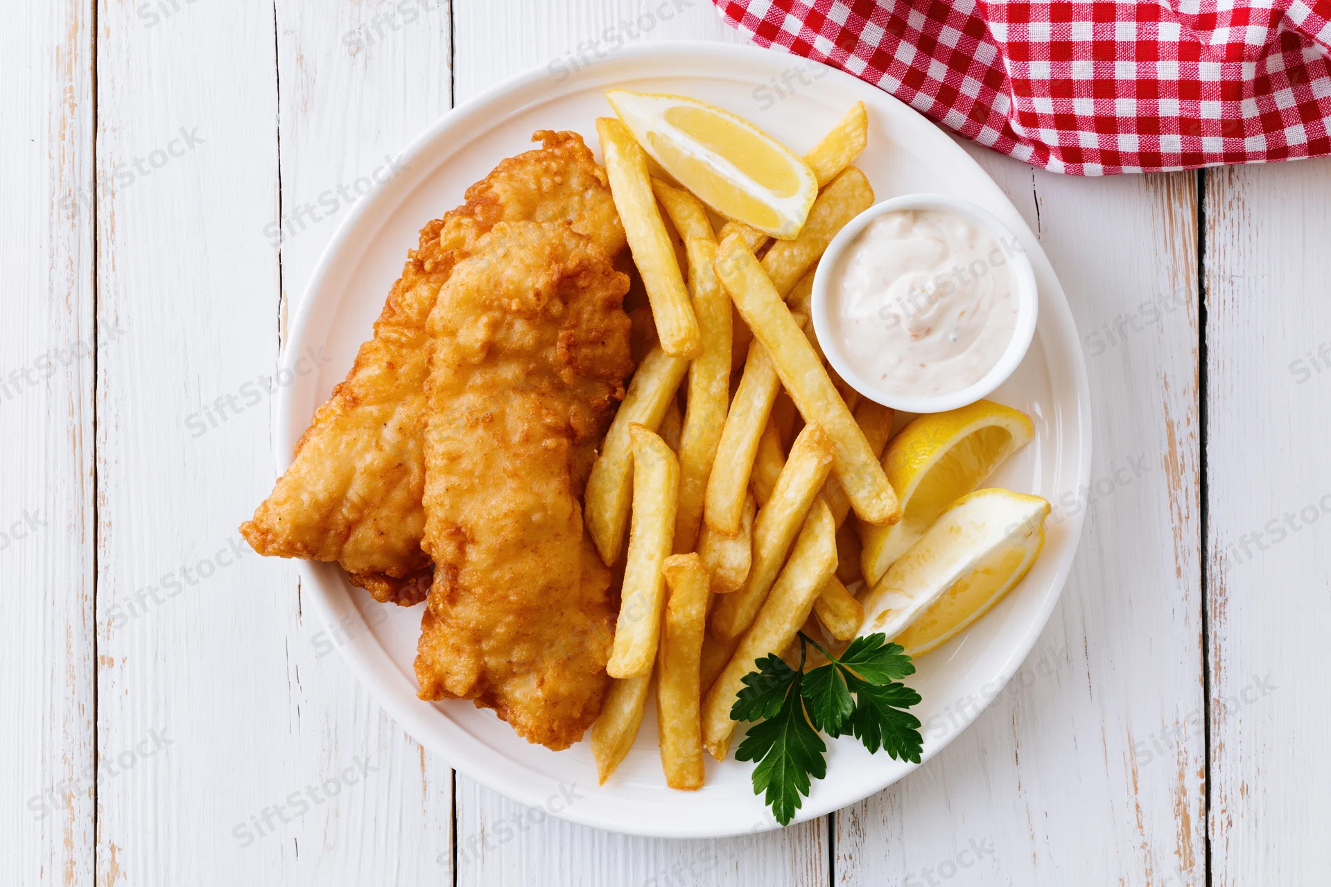 Fried fish and french fries with tartar sauce and lemon wedges on a white plate over a wooden table