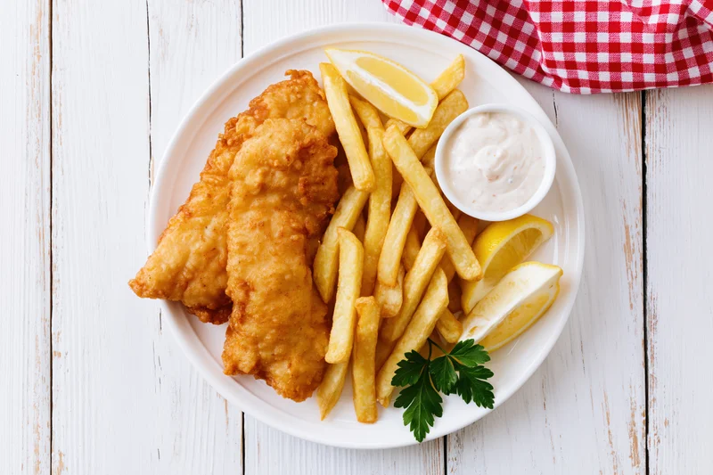Fried fish and french fries with tartar sauce and lemon wedges on a white plate over a wooden table