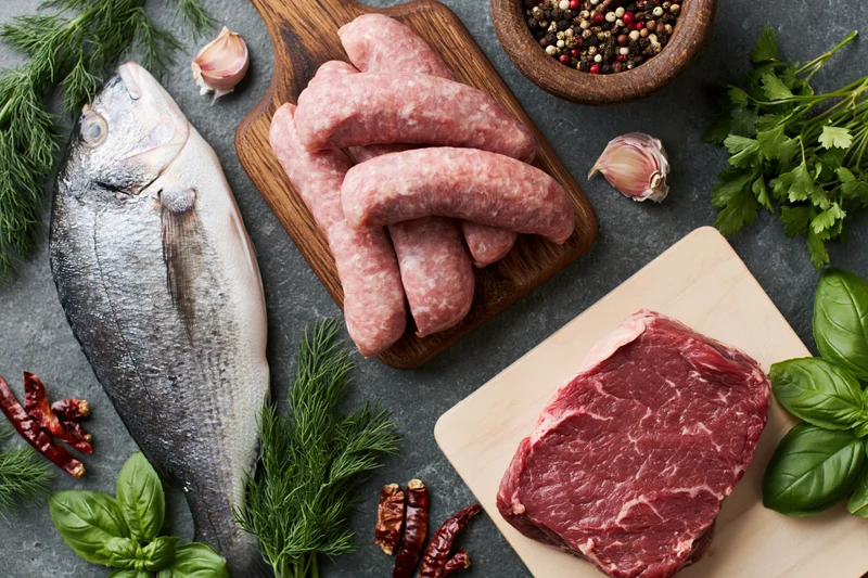 Raw fish, sausage and beef arranged with fresh herbs and spices on dark countertop