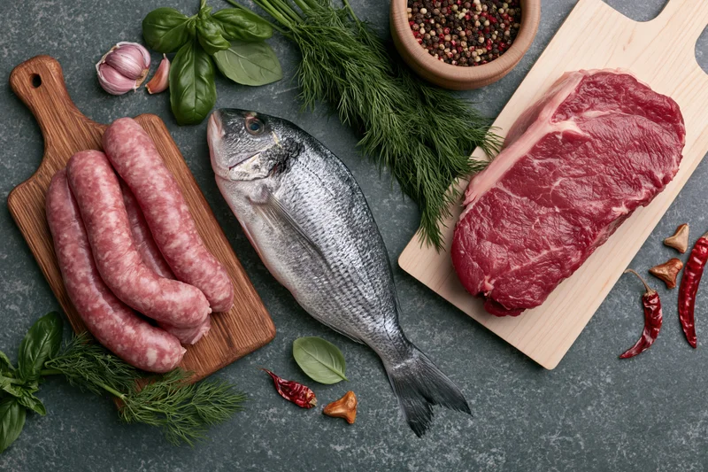 Raw meat, fish and sausage arranged with fresh herbs on dark countertop