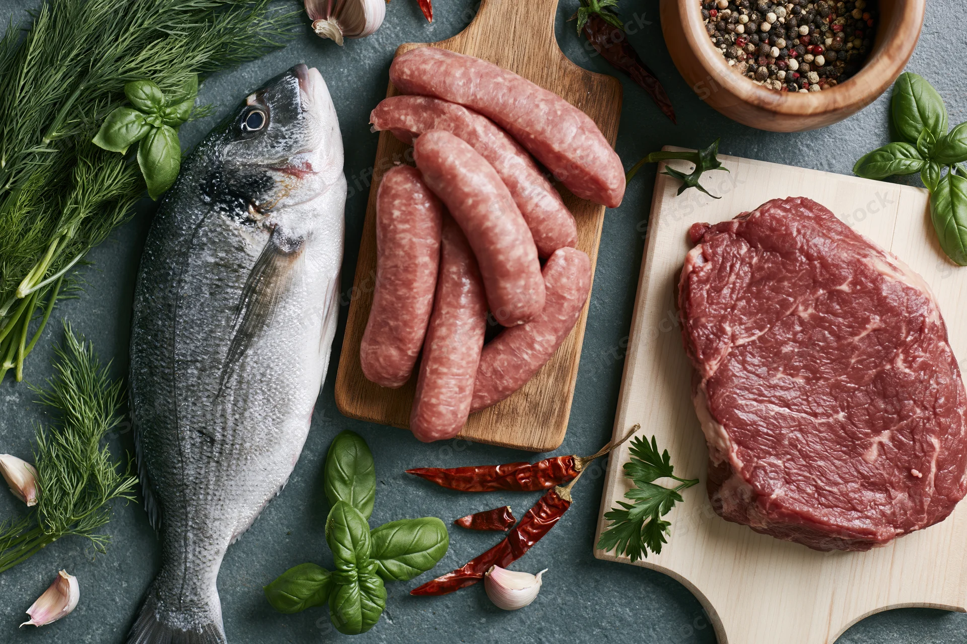 Raw fish, sausage, and steak arranged with fresh herbs on dark countertop