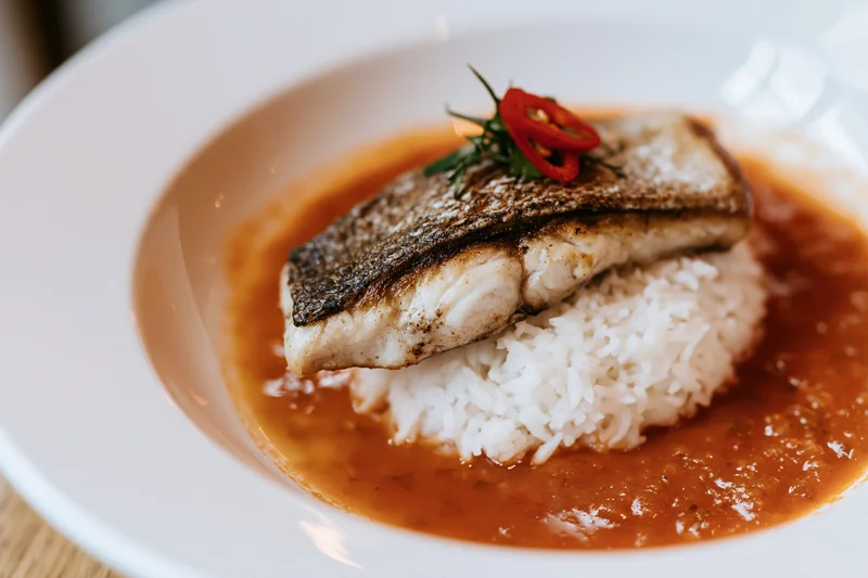 Pan-seared fish fillet with crispy skin on a mound of white rice in tomato sauce on a white plate