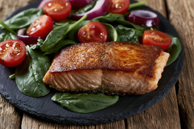 Pan-seared salmon fillet with crispy golden-brown skin served on a bed of spinach and cherry tomatoes on a slate plate