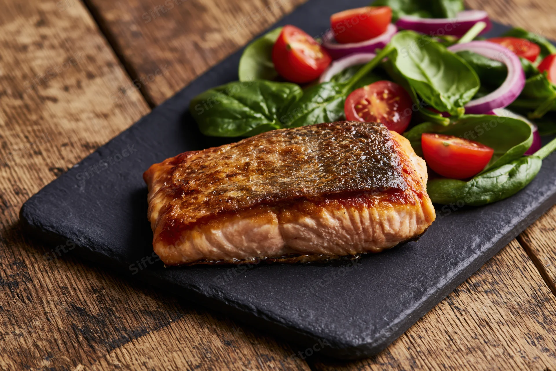 Pan-seared salmon fillet with crispy skin served on slate plate with spinach and tomato salad on wooden table