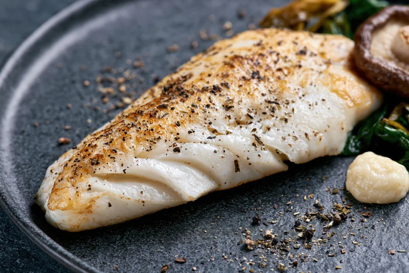 Pan-seared white fish fillet on dark matte ceramic plate with herbs and side vegetables