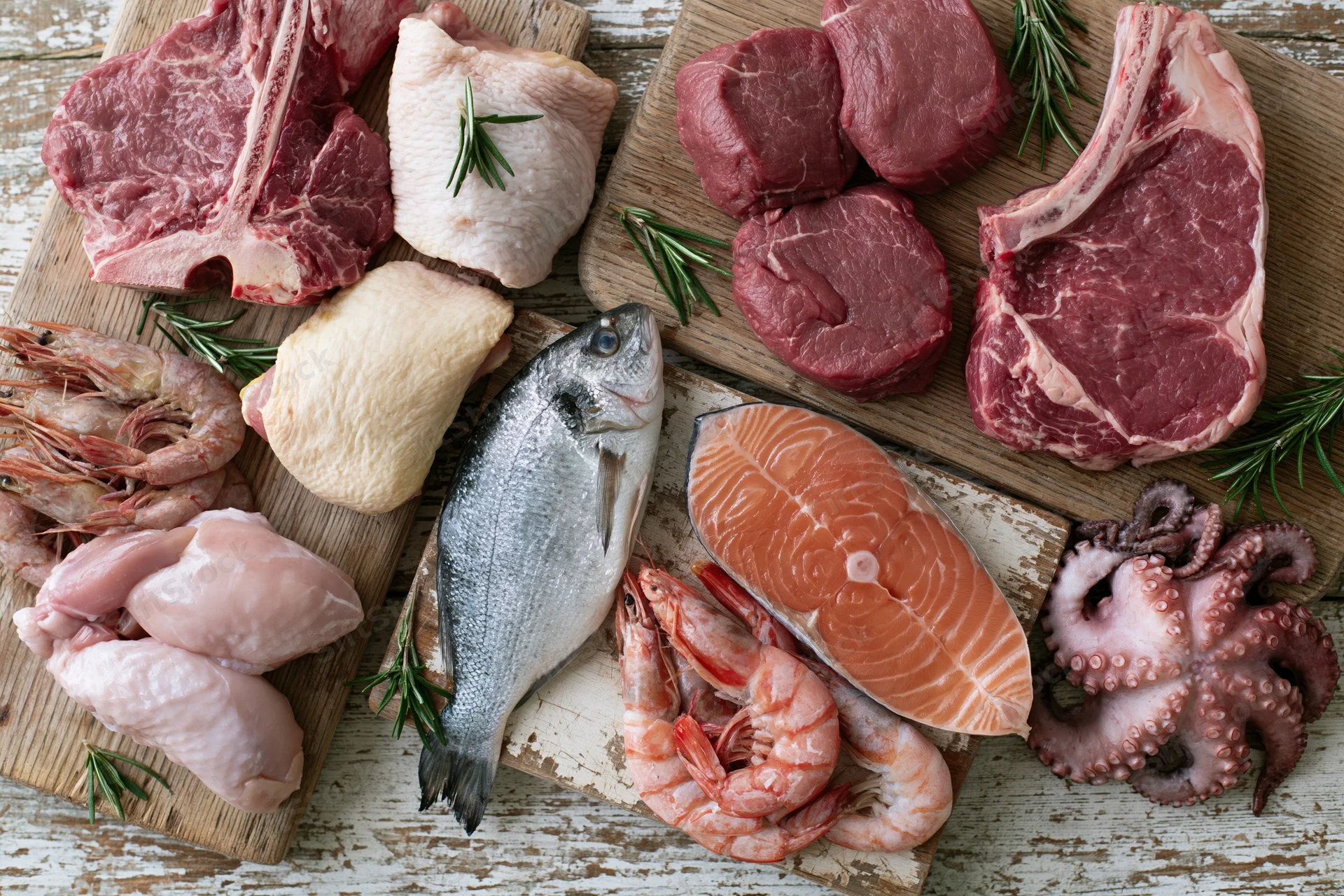 Raw meat seafood and poultry arranged on wooden cutting boards with rosemary sprigs