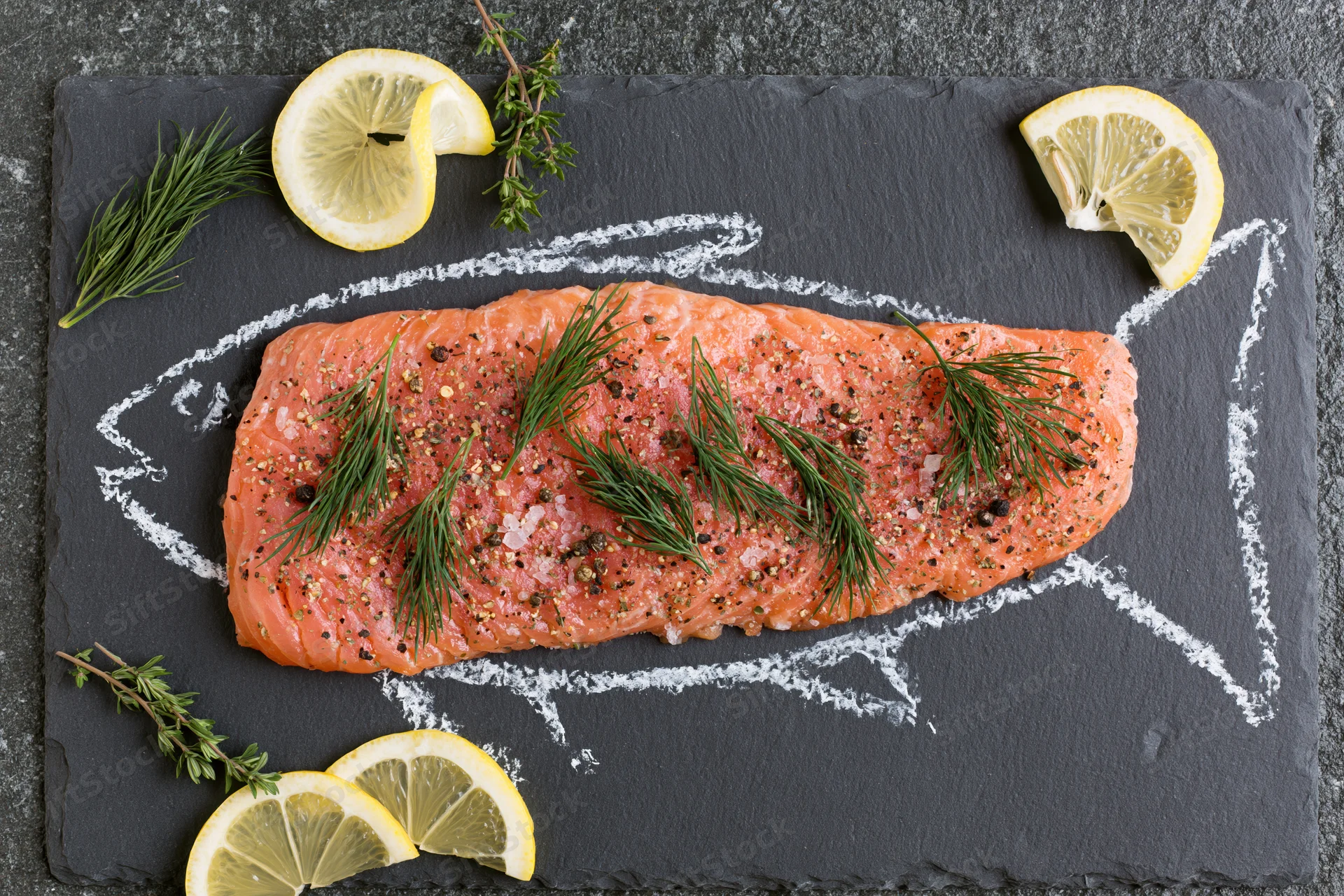 Raw salmon fillet seasoned with dill and pepper on slate plate with lemon slices and herbs arranged in fish outline